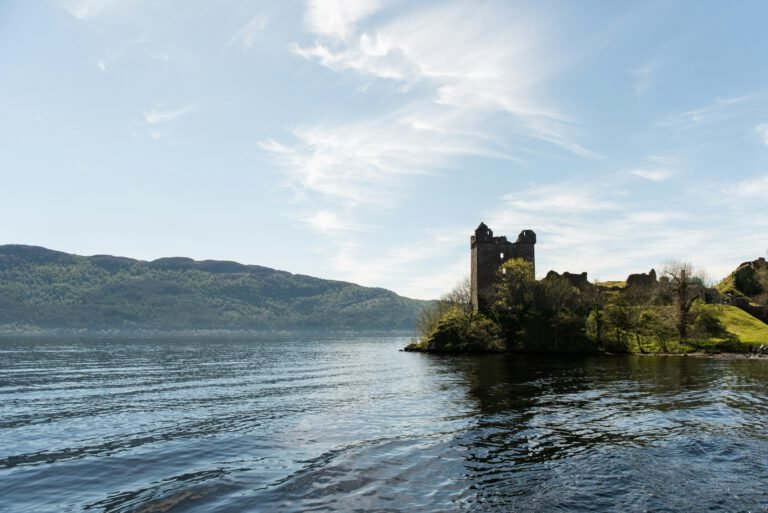 The ruins of Urquhart Castle on the shore of a calm Loch Ness under a bright blue sky with wispy clouds.