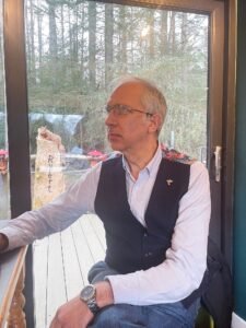 Scottish poet Sam Steele wearing a dark waistcoat with floral shoulder detailing, looking thoughtfully out a window toward a woodland garden.