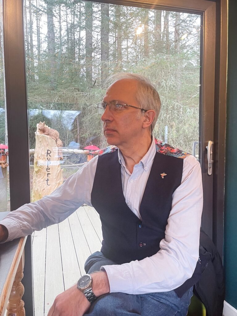Scottish poet Sam Steele wearing a dark waistcoat with floral shoulder detailing, looking thoughtfully out a window toward a woodland garden.
